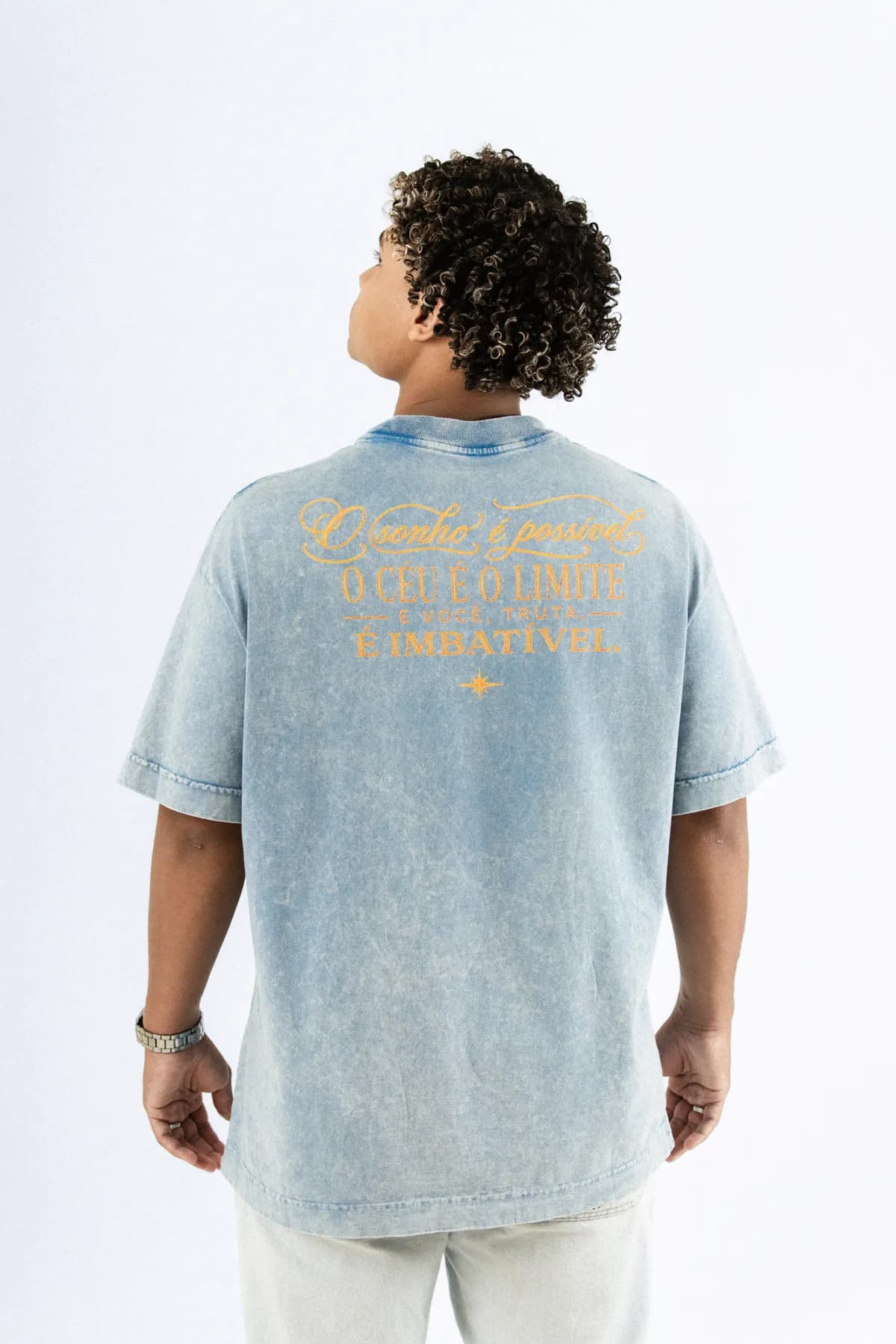 Modelo de costas, com a camiseta azul estonada e a frase bordada em dourado nas costas.