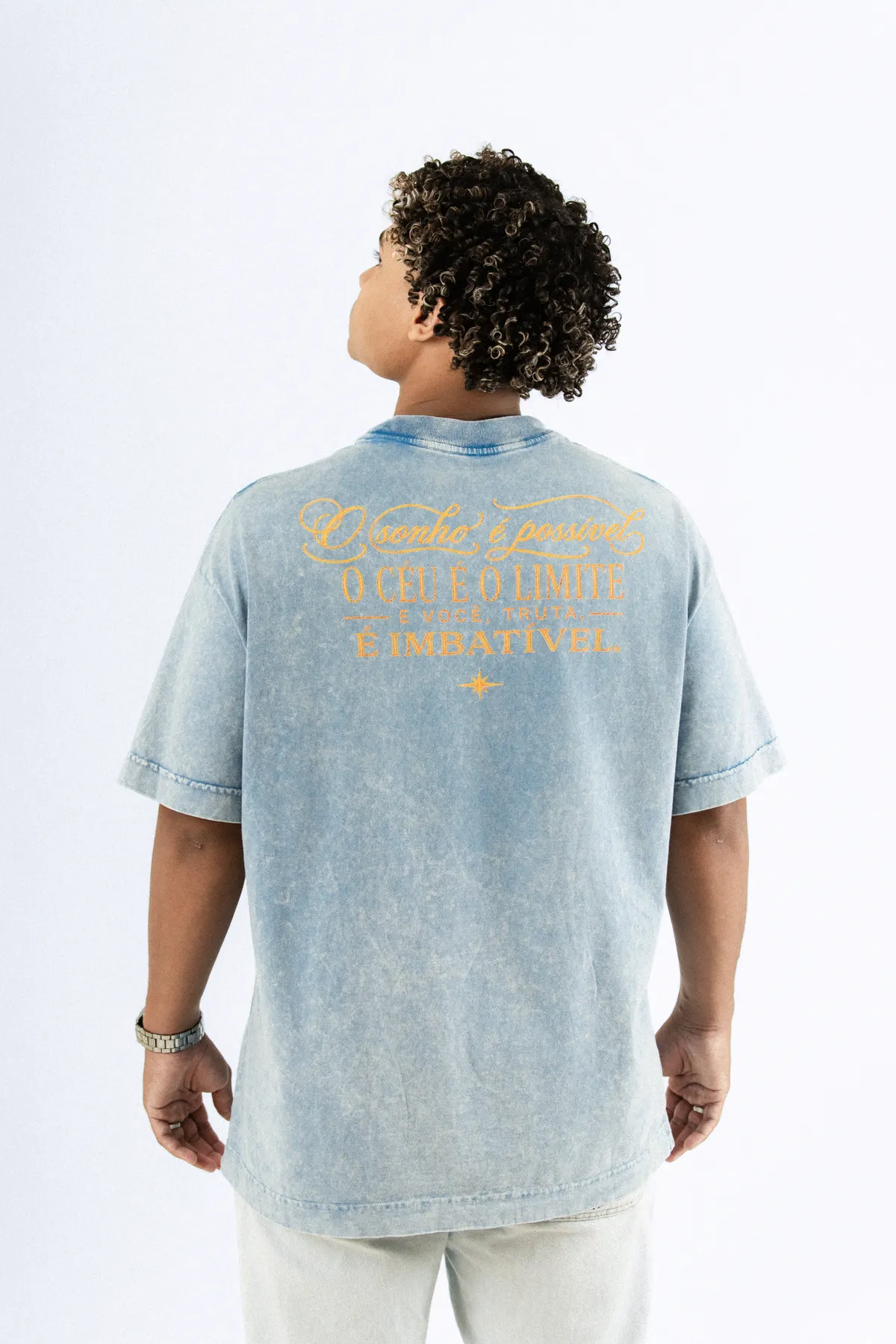 Modelo de costas, com a camiseta azul estonada e a frase bordada em dourado nas costas.