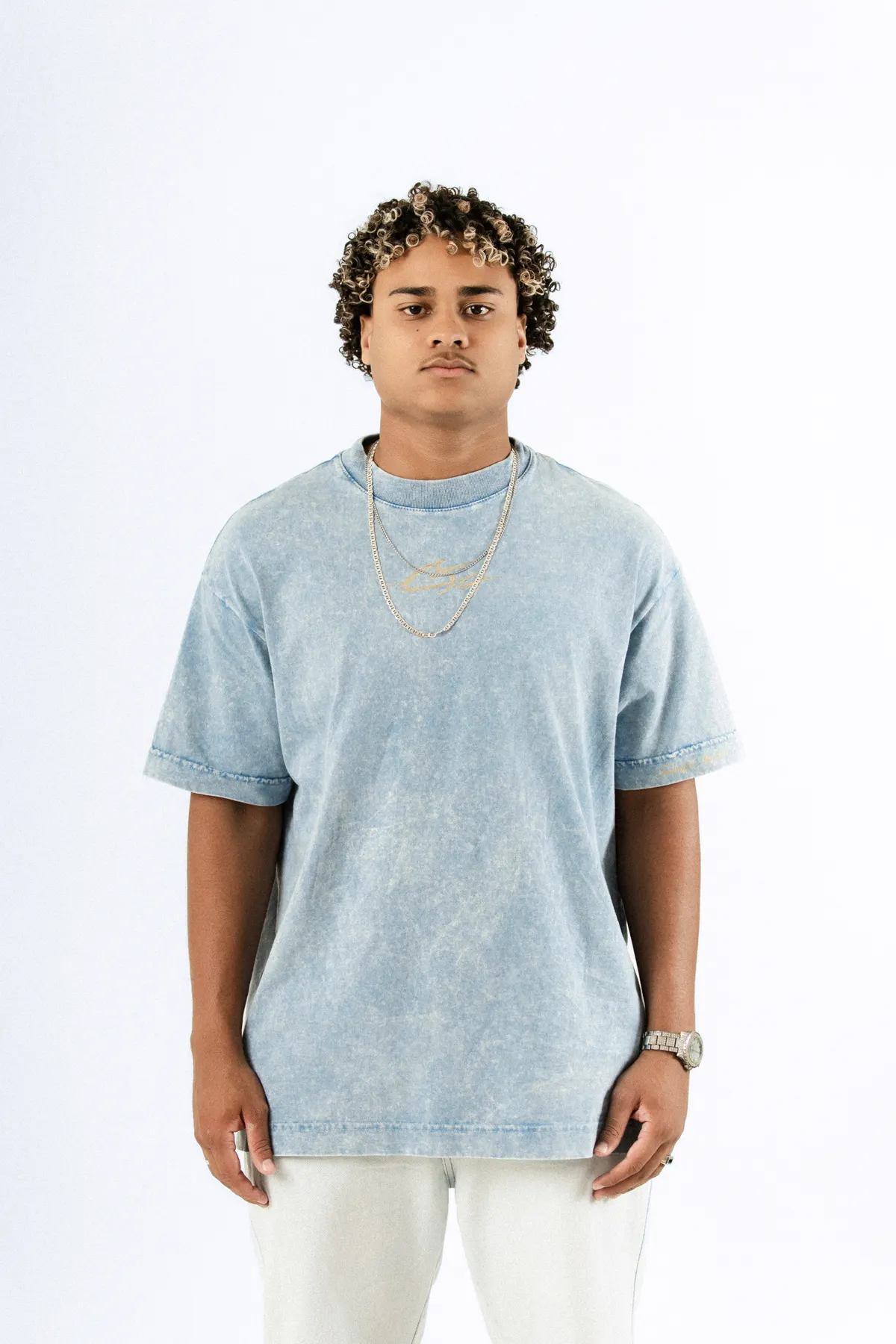 Modelo de cabelo cacheado, de frente, com a camiseta azul estonada e corrente prateada.